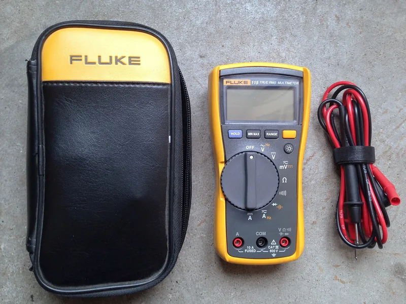 Fluke 115 digital multimeter in its yellow holster, with the black carrying case and coiled test leads, on a concrete floor.
