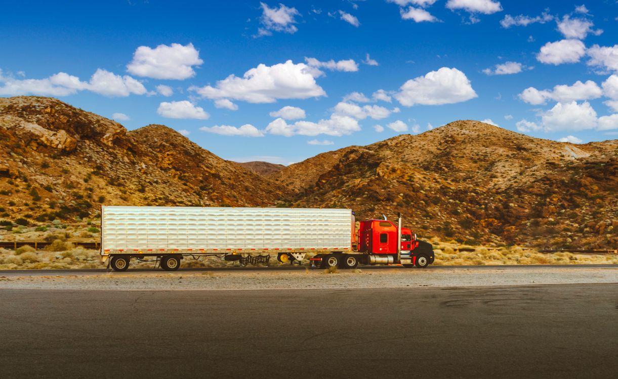 Red semi truck with a dry van trailer parked along a desert highway under a blue sky.